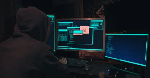 Hacker in a hood sits in front of computer screens and hacks databases Stock Photos