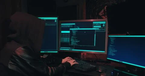 Hacker in a hood sits in front of computer screens and hacks databases Stock Photos