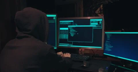 Hacker in a hood sits in front of computer screens and hacks databases Stock Photos