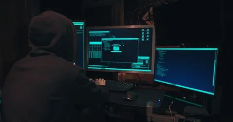 Hacker in a hood sits in front of computer screens and hacks databases Stock Photos