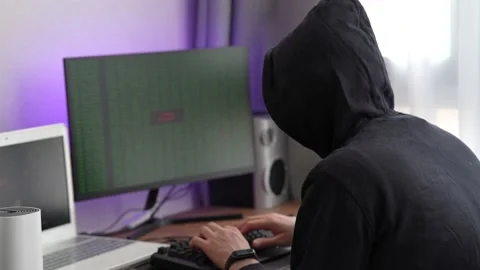 A hacker in a hood types program code. Young man criminal wearing black hoodie Stock Footage 179444422