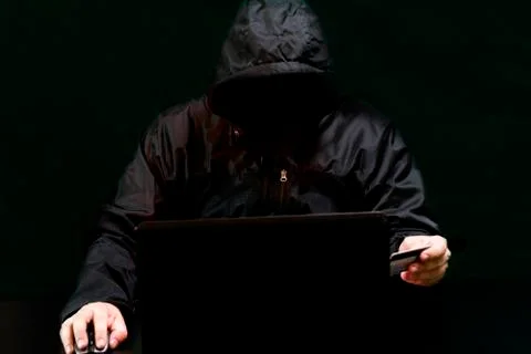 A hacker in a hood, without a face, on a black background sits at a laptop and 스톡 사진