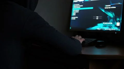 A hacker in a hooded sweatshirt is sitting at a computer in the dark. light from Stock Footage 167156773