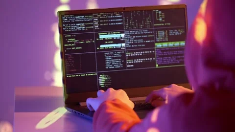 A hacker in a hooded sweatshirt writes code on a laptop terminal with system Stock Footage 307010300