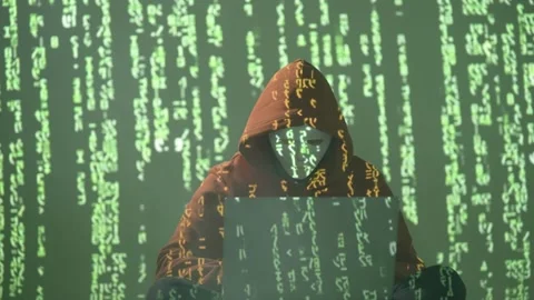 Hacker in Mask and Hoodie Using Laptop Surrounded by Falling Green Code in 스톡 동영상 307179275