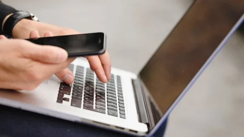 Hacker studies info in phone types on laptop put on knees Stock Footage 111611481