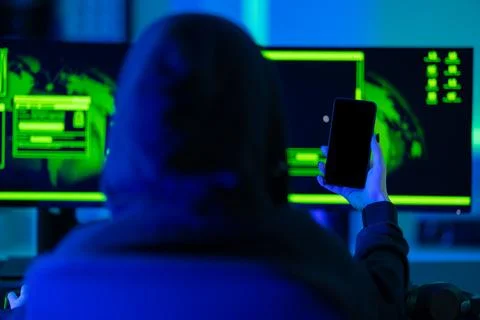 Hacker using smartphone in front of dual monitors with green glow Stock-Fotos