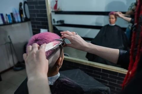 Hairdresser cuts Asian woman with short pink hair in barbershop. Stock Photos