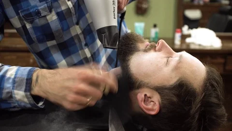 Hairstyling process. Close-up of a barber drying hair of a young bearded man Stock Footage 69990648