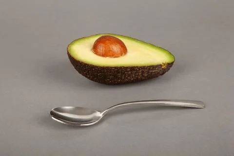 A half of an avocado with a bone and a spoon lie on a gray background. Stock Photos