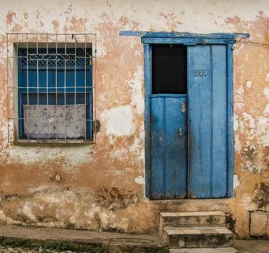Half-open blue door plus window with grate on tattered yellow wall with stone Foto stock