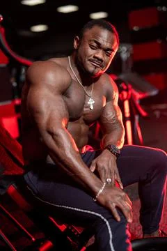 Halfnaked focused african american bodybuilder sitting in modern dark gym wit Stock Photos