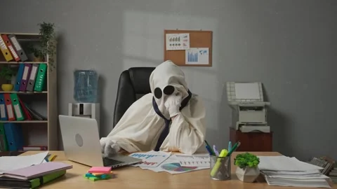 Halloween businessman ghost in white sheet sits at office desk works on laptop Stock Footage 313387003
