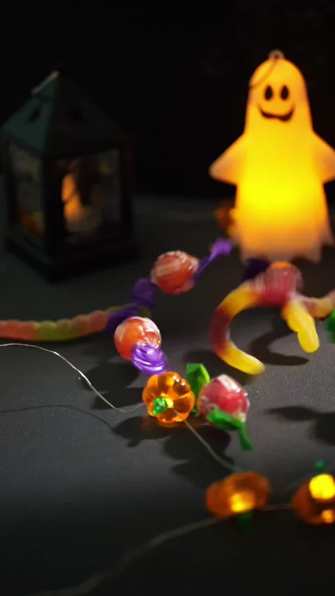 Halloween candy falling on dark table by glowing ghost lamp Stock Footage 343569910
