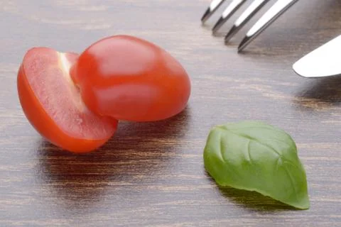 Halved cherry tomatoes and basil leaves on a dark table. Fork and knife. Stock Photos