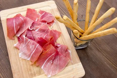 Ham on cutting board with bread sticks on wooden table seen from above Stock Photos