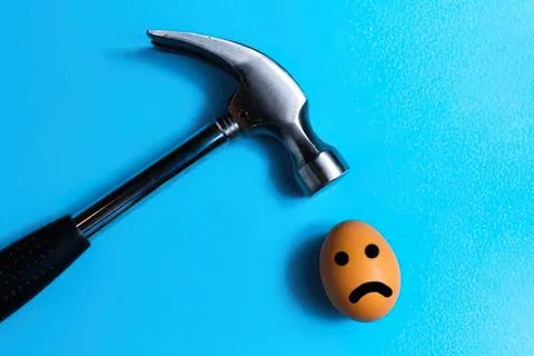 Hammer hitting egg with sad face on blue background Stock Photos