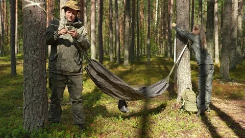 A hammock is being hung between trees by a man and a woman. In the forest, a Stockbeeldmateriaal 318659716