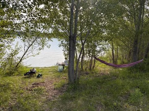 Hammock strung between trees in a peaceful waterside forest clearing with campin Stock Photos