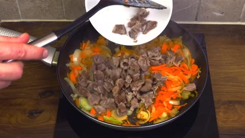 Hand adding chicken gizzards to carrots and onions in pan Stock Footage 345422264
