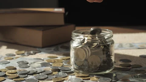 Hand Adding Coin Into Savings Jar With Cash and Books Background Stock Footage 346035152