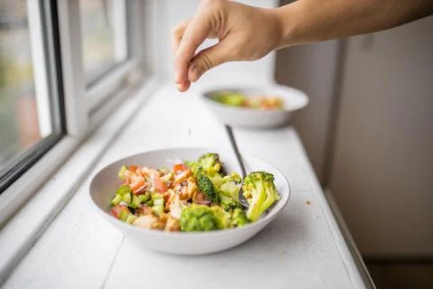 Hand adding a pinch of salt to a broccoli and chicken salad next to a window Stock Photos