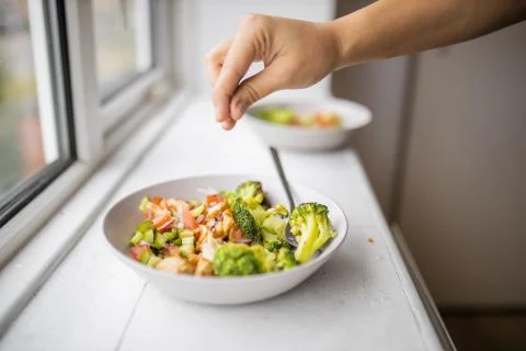 Hand adding a pinch of salt to a broccoli and chicken salad next to a window Stock Photos