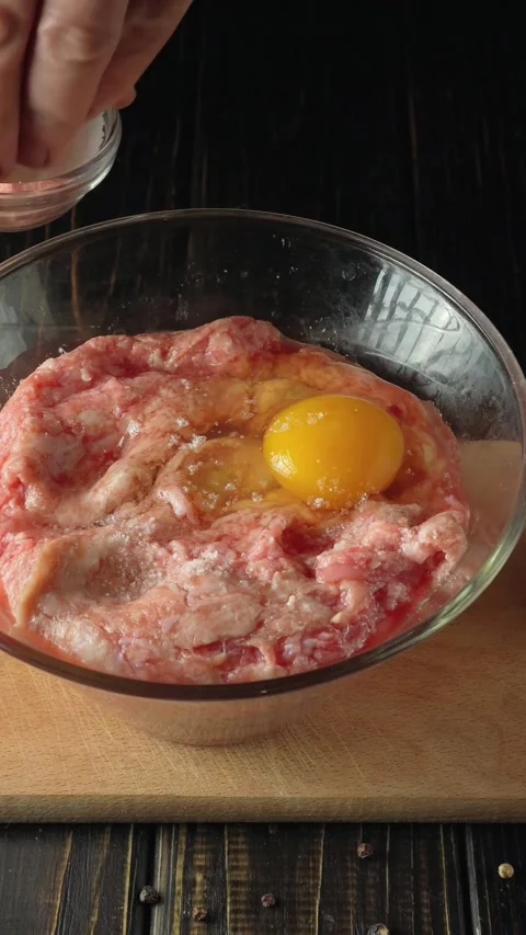 Hand adding salt to raw meat mixture with egg yolk in glass bowl on wooden Stock Footage 339320484