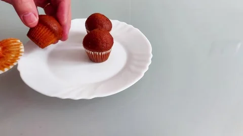 Hand Adding Third Brown Muffin to Plate Beside Two on Table Surface 스톡 동영상 328545409