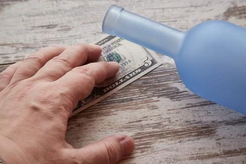 A hand and five dollars next to a bottle on a worn table, problems with alcoh Stock Photos