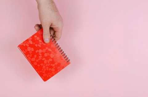Hand and notepad on pink backdrop. An adult man holds a closed red notebook o Stock Photos