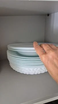 A hand arranges white plates on a kitchen shelf during domestic chores. Stock Photos