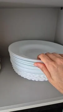 A hand arranges white plates on a kitchen shelf during domestic chores. Stock Photos