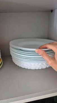 A hand arranges white plates on a kitchen shelf during domestic chores. Stock Photos
