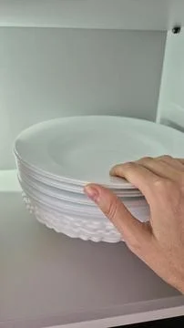 A hand arranges white plates on a kitchen shelf during domestic chores. Stock Photos