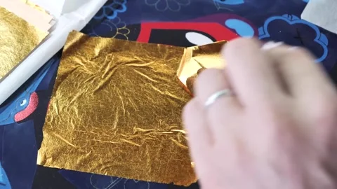 Hand carefully presses a square of gold leaf onto navy fabric during a textile Vídeos de archivo 339123569