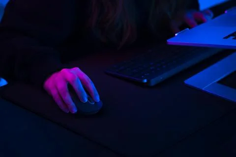 Hand on a computer mouse at a dark desk lit by pink and blue light Stock Photos