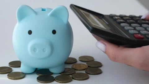 Hand counting on calculator next to piggy bank and coins on white table Video stock 312807548
