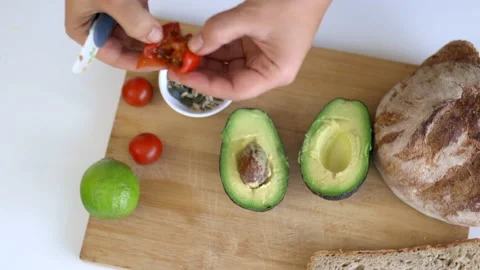 Hand cutting tomato in half above cutting board with sliced bread and avocados 스톡 동영상 144925582