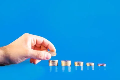 Hand depositing a euro coin in a stack of coins on blue background. Business  Stock Photos