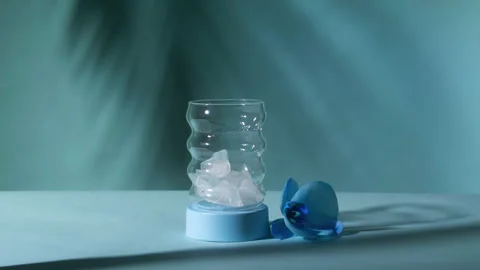 Hand Dropping Ice Cubes into Empty Bubble Glass Next to Blue Orchid Stock Footage 334264206