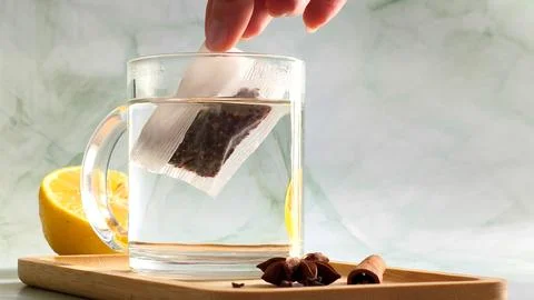 Hand dropping tea bag into clear glass cup with boiling water Stock Photos