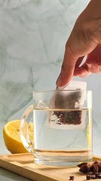 Hand dropping tea bag into clear glass cup with boiling water Stock Photos