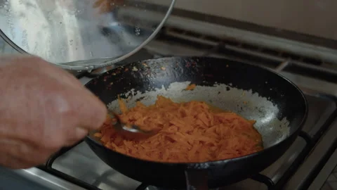 The hand of an elderly man stirs finely grated carrots fried in oil in a pan 스톡 동영상 337121203