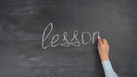 The hand of a female teacher writes the word LESSON on a black chalk board Stock Footage