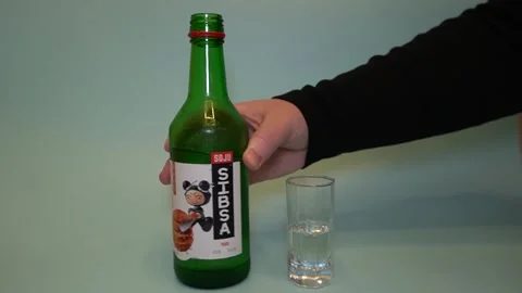 A hand with a filled glass brings it to the neck of a green bottle of soju. Stock Footage 320145942