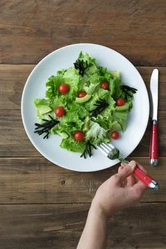 Hand with fork eating assorted salad on white plate on wooden background. Stock Photos
