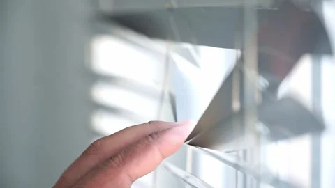 A hand gently opens window blinds, allowing natural sunlight into a modern home Stock Footage 340323302