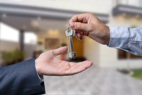 A hand giving a key to another hand. House in the background. Stock Photos