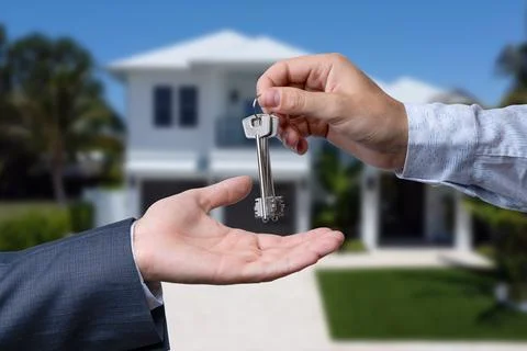 A hand giving a key to another hand. House in the background. Stock Photos